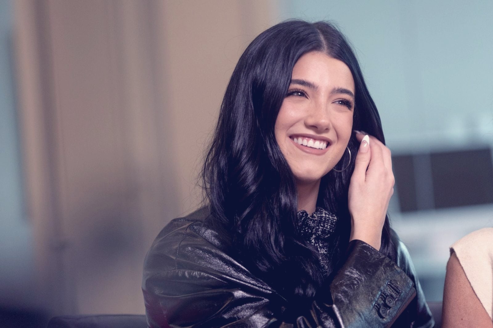 Charli D'Amelio smiling, one hand in her long dark hair, in a black leather jacket, seated indoors in an interview setting