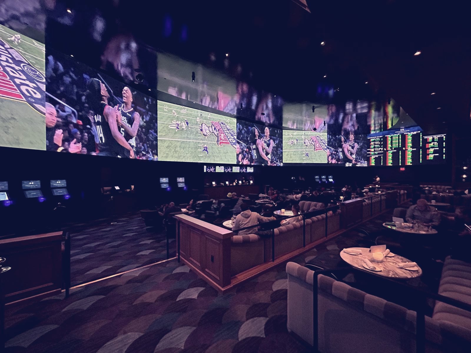 Context image: the sportsbook at Fontainebleau Las Vegas, a wall of video screens and an odds board above lounge seating and gaming kiosks, with a few patrons watching. Photographed in January 2024.