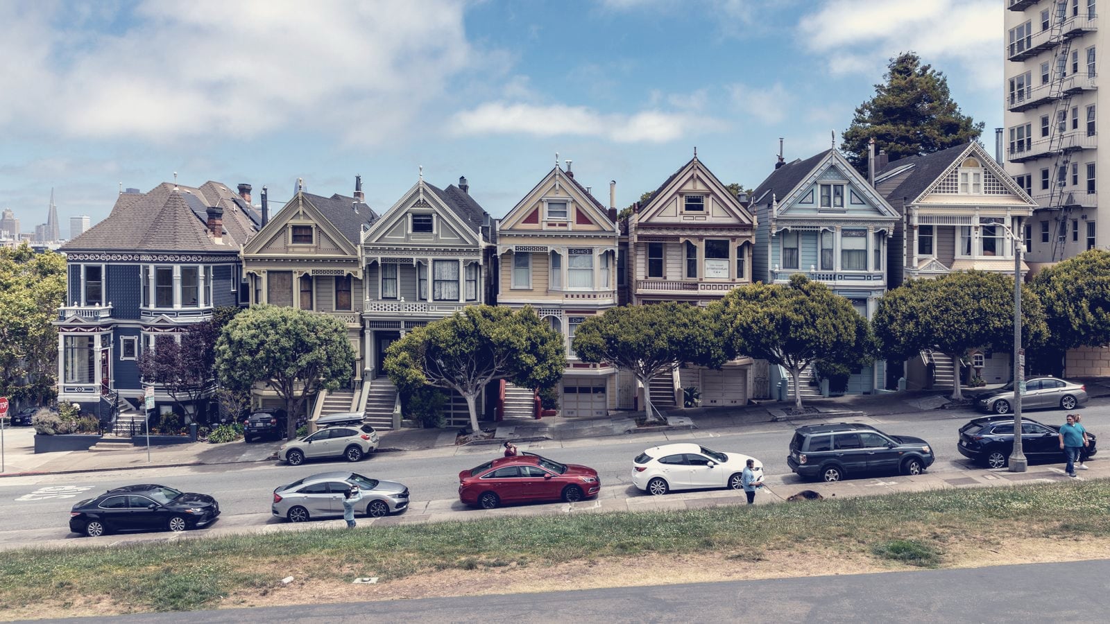 Context image: a row of Victorian houses facing Alamo Square in San Francisco, the park that hacker-house mansions overlook, photographed in July 2022 under a partly cloudy sky with parked cars along the street.