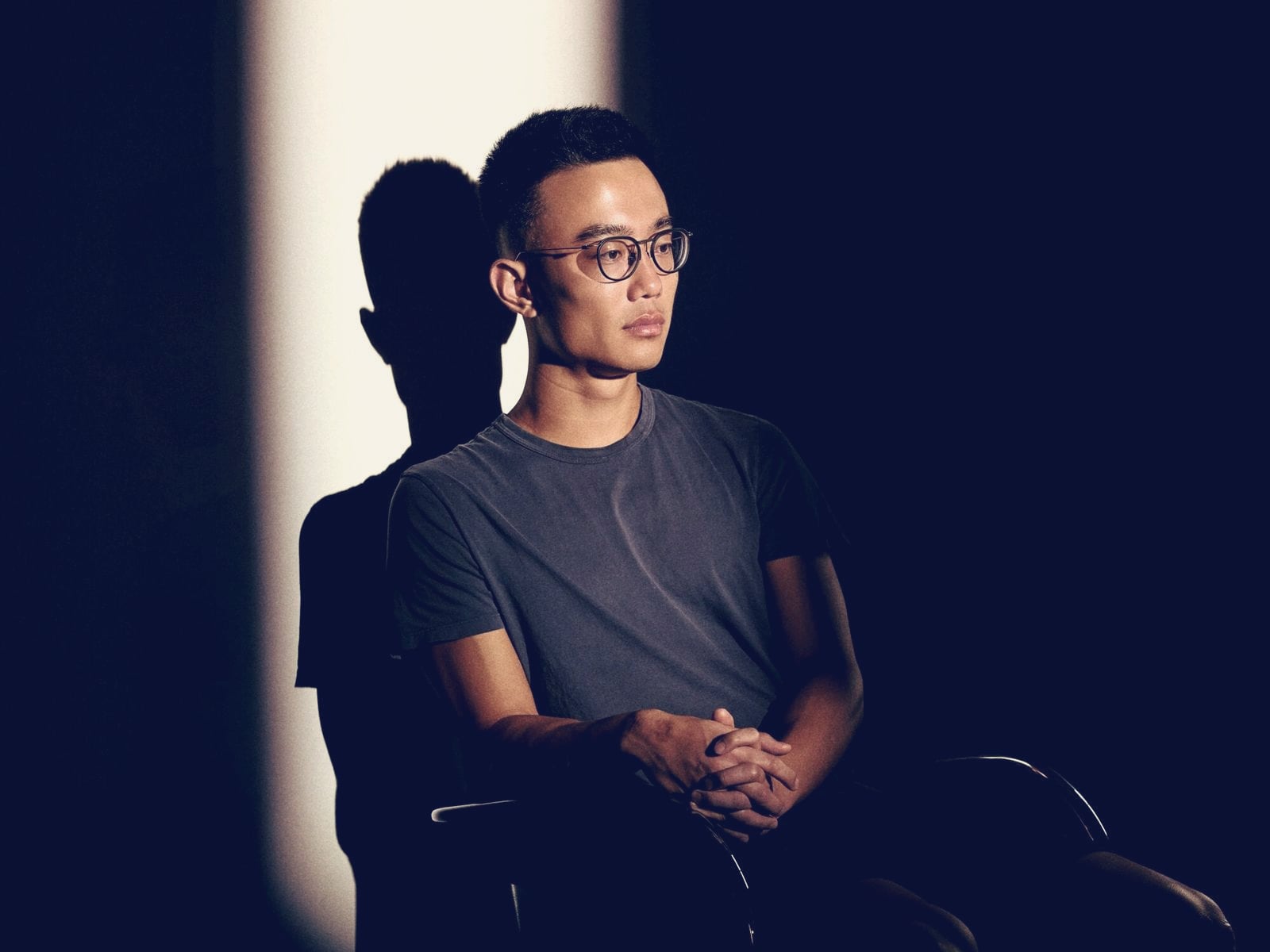 Jeff Yan, founder of Hyperliquid, seated in a pool of light against a dark studio backdrop, his shadow cast on the wall behind him.