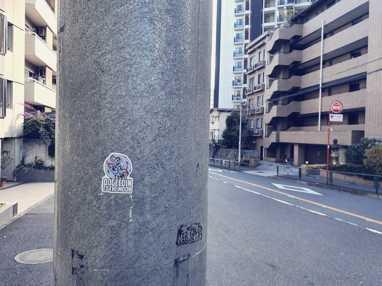 A context image, not of the events described: a small Dogecoin 'to the moon' sticker, showing an astronaut dog on a rocket, stuck to a metal utility pole on a residential street in Tokyo.