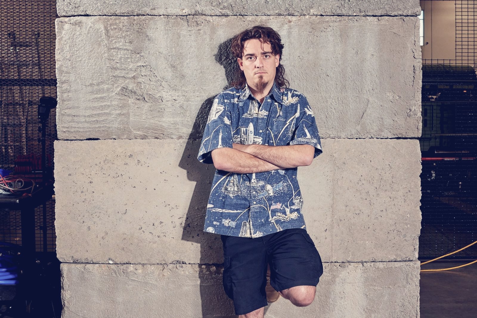 Palmer Luckey leaning against a concrete wall at Anduril's factory in Santa Ana, California, arms crossed, in a space-themed shirt