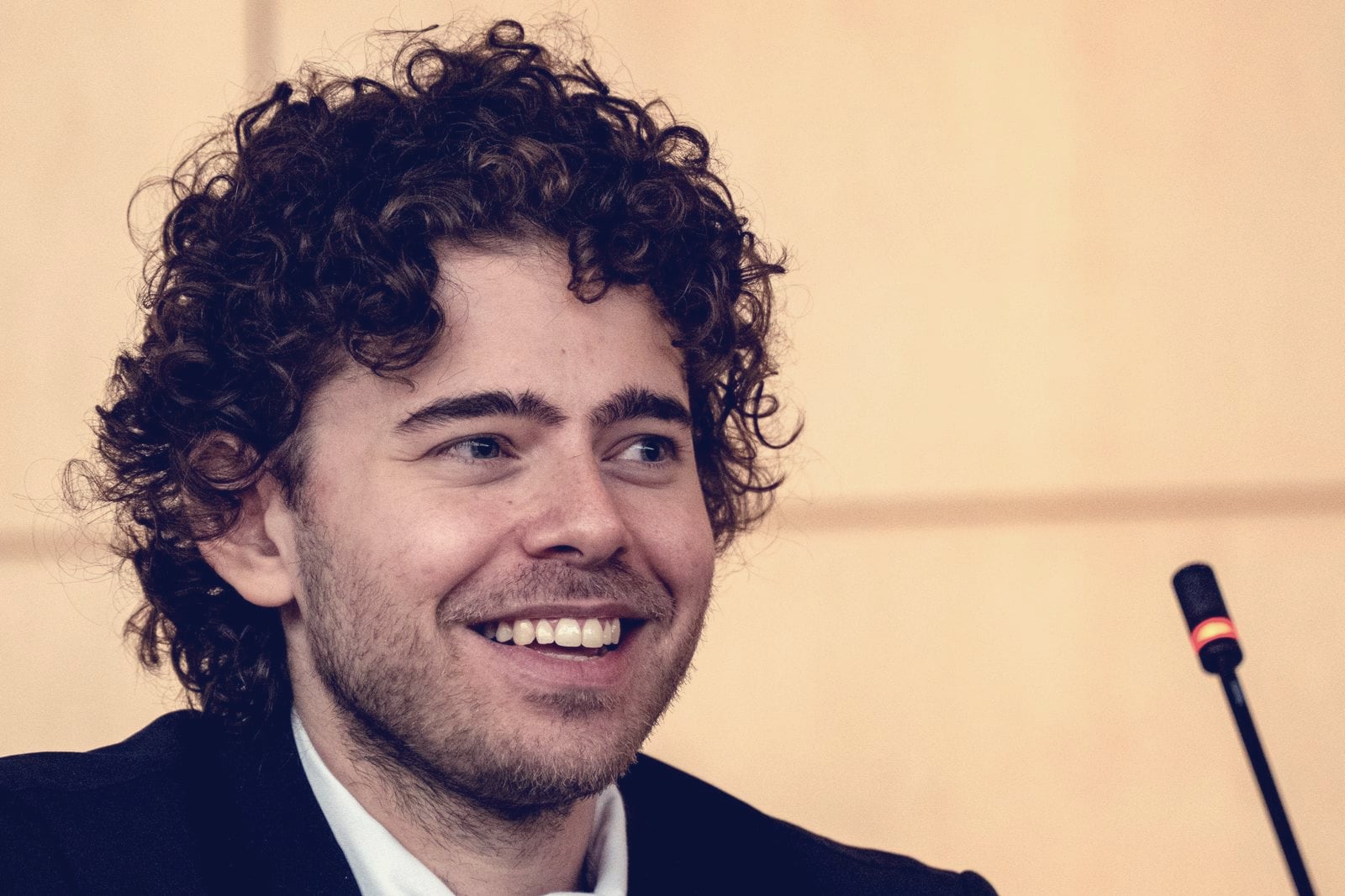 Shayne Coplan, founder of Polymarket, smiling mid-sentence beside a microphone against a warm beige wall.