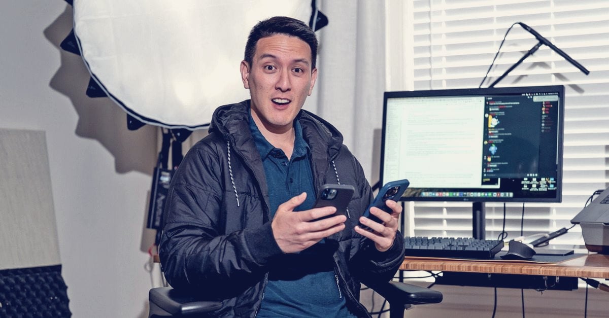 Humphrey Yang, the personal finance creator, sits in his San Francisco studio holding two phones, with a softbox light behind him and a monitor showing a document and a dark chat sidebar on his desk.
