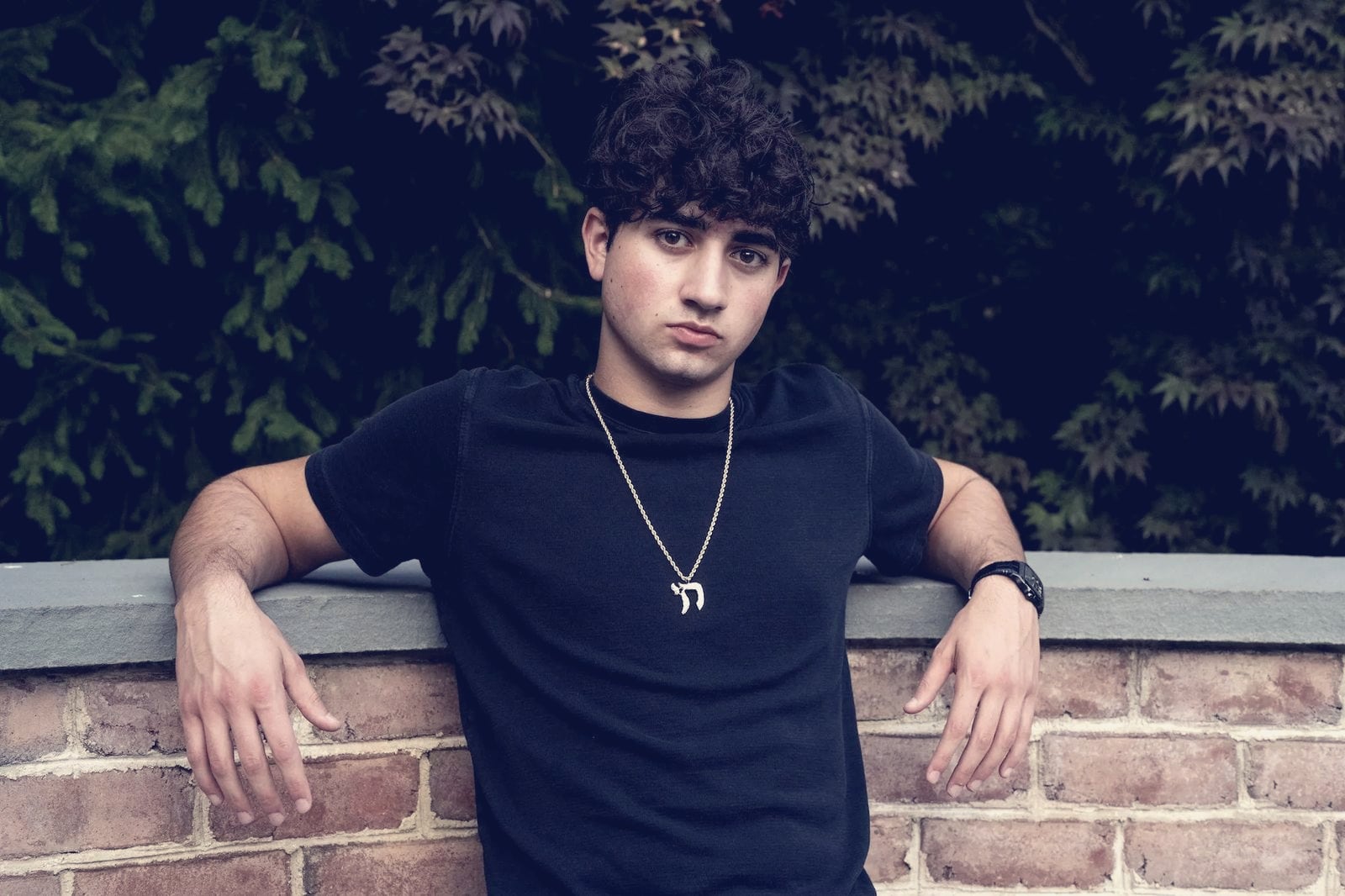 Zach Yadegari, co-founder of Cal AI, in a black T-shirt and gold chain leaning on a low brick wall in front of dark foliage, in a portrait supplied by MyFitnessPal.
