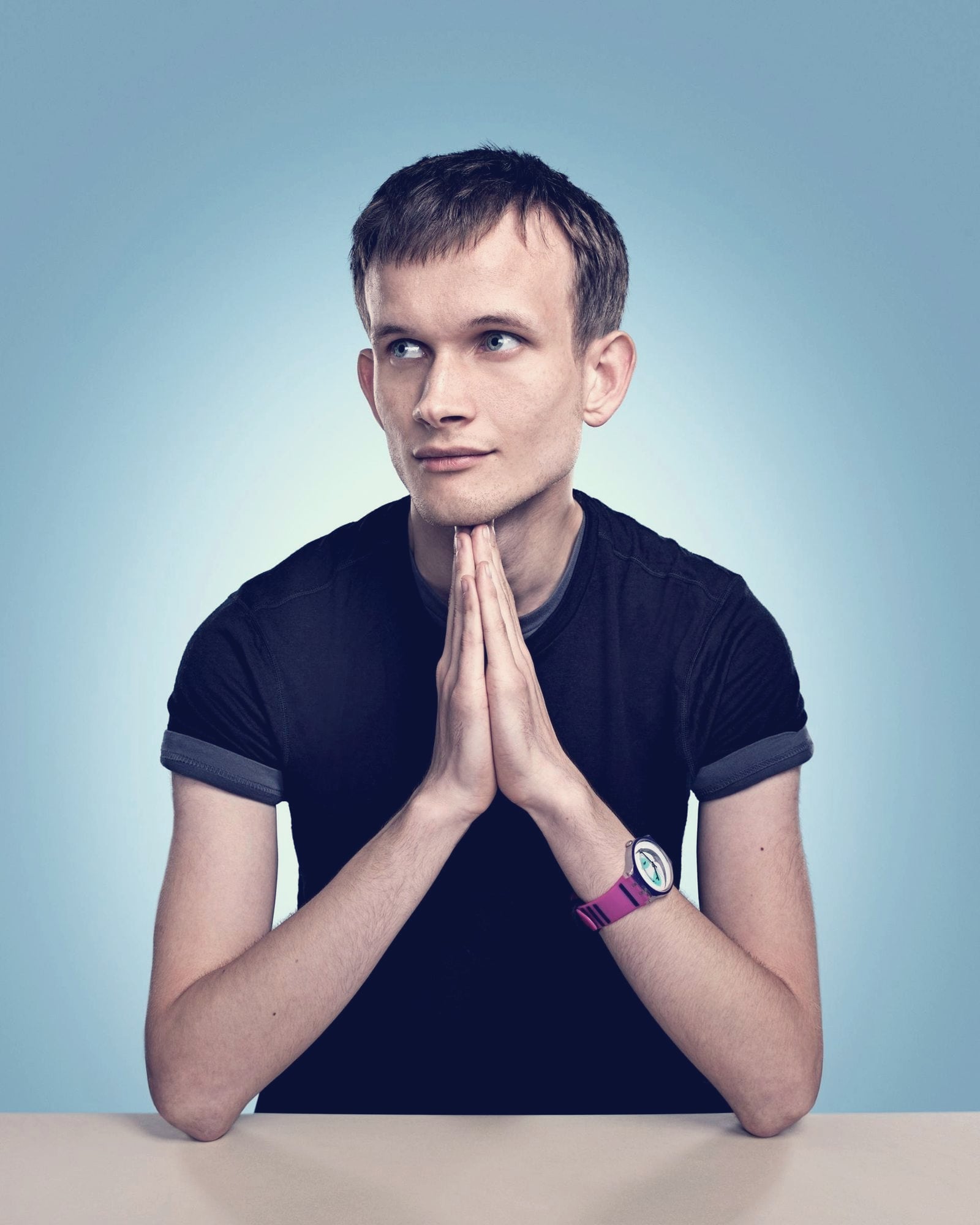 Vitalik Buterin seated at a table against a blue backdrop, hands pressed together under his chin, glancing to the side